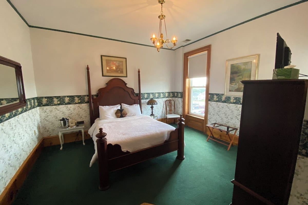 A bedroom with a four-poster bed, a chandelier, a dresser, and a window with a view.