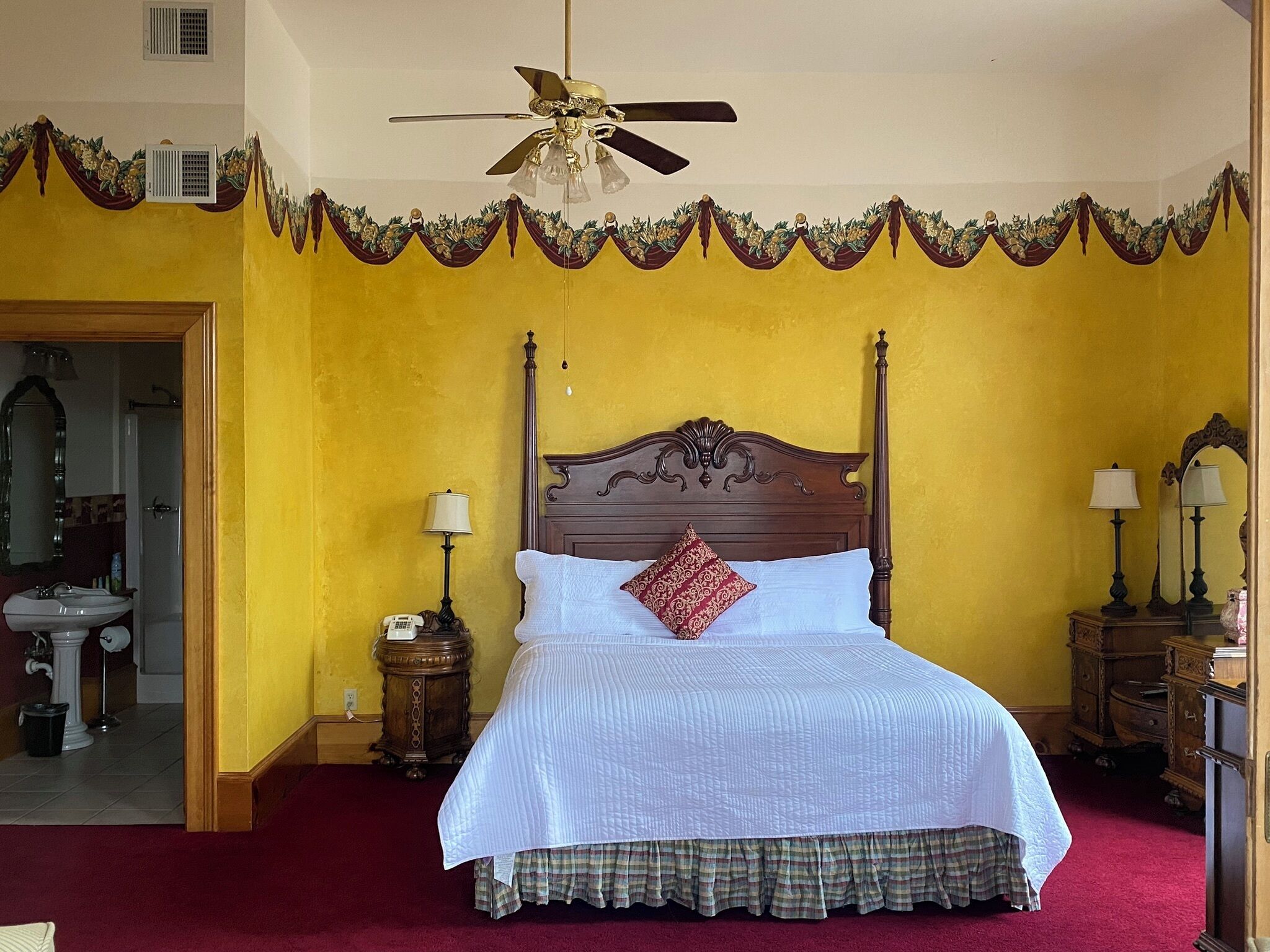 Un dormitorio con cama de madera, ventilador de techo, baño con lavamanos y espejo, y alfombra roja.