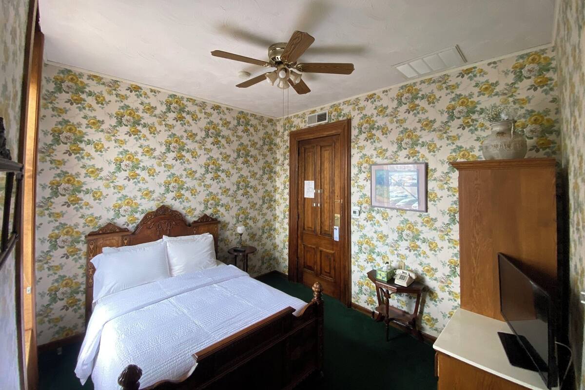 A bedroom with floral wallpaper, a wooden bed, a ceiling fan, a wooden wardrobe, and a desk with a chair.