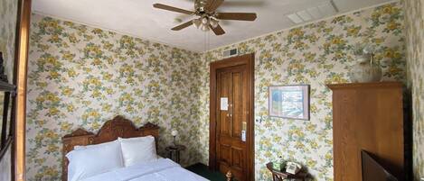 A bedroom with floral wallpaper, a wooden bed, a ceiling fan, a wooden wardrobe, and a desk with a chair.