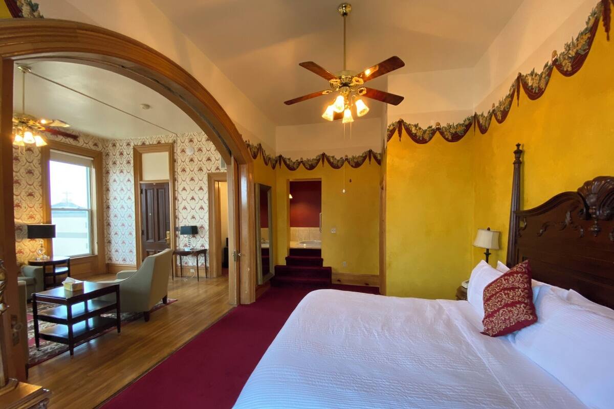 A hotel room with a large bed, a desk with a chair, a mirror, and a ceiling fan.