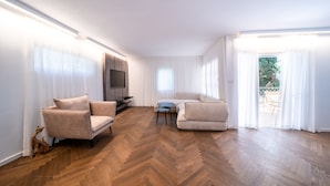Uma sala de estar ampla com piso de madeira em padrão herringbone, dois sofás bege, uma mesinha azul e uma TV fixada na parede.
