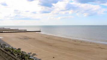 A wide beach with a calm sea, a wooden pier, and a stone wall with rocks.