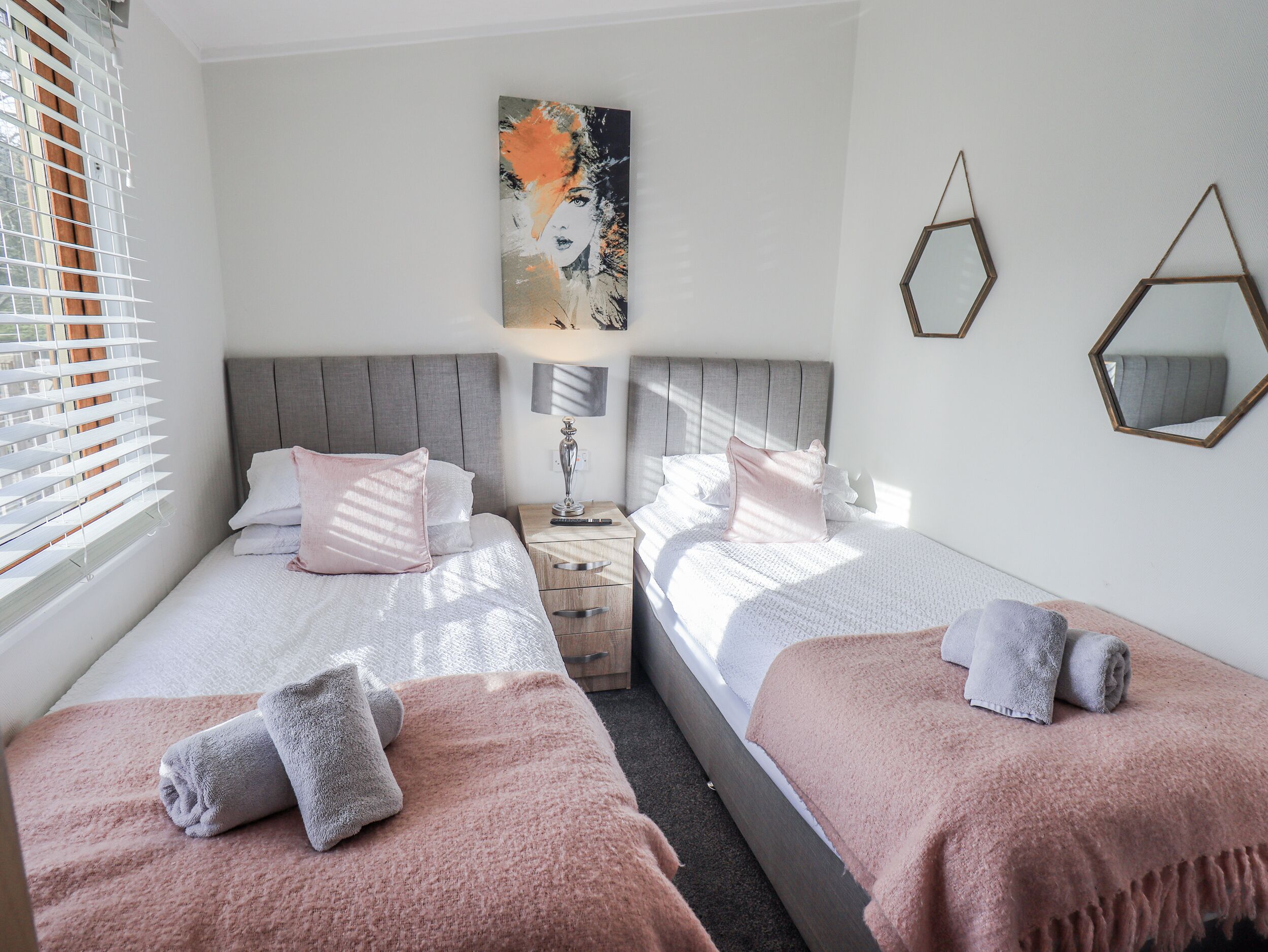 A hotel room with two beds, each with a pink blanket and a rolled towel. There is a painting on the wall and two hexagonal mirrors.