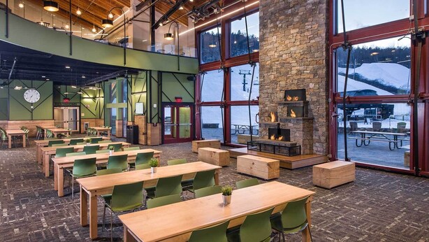 A spacious indoor area with multiple wooden tables and green chairs, large windows offering a view of a snowy mountain, and a stone fireplace with a fire. - Modern Mount Snow Gnarvana - Balcony, Loft, Pets, Hiking (Dover)