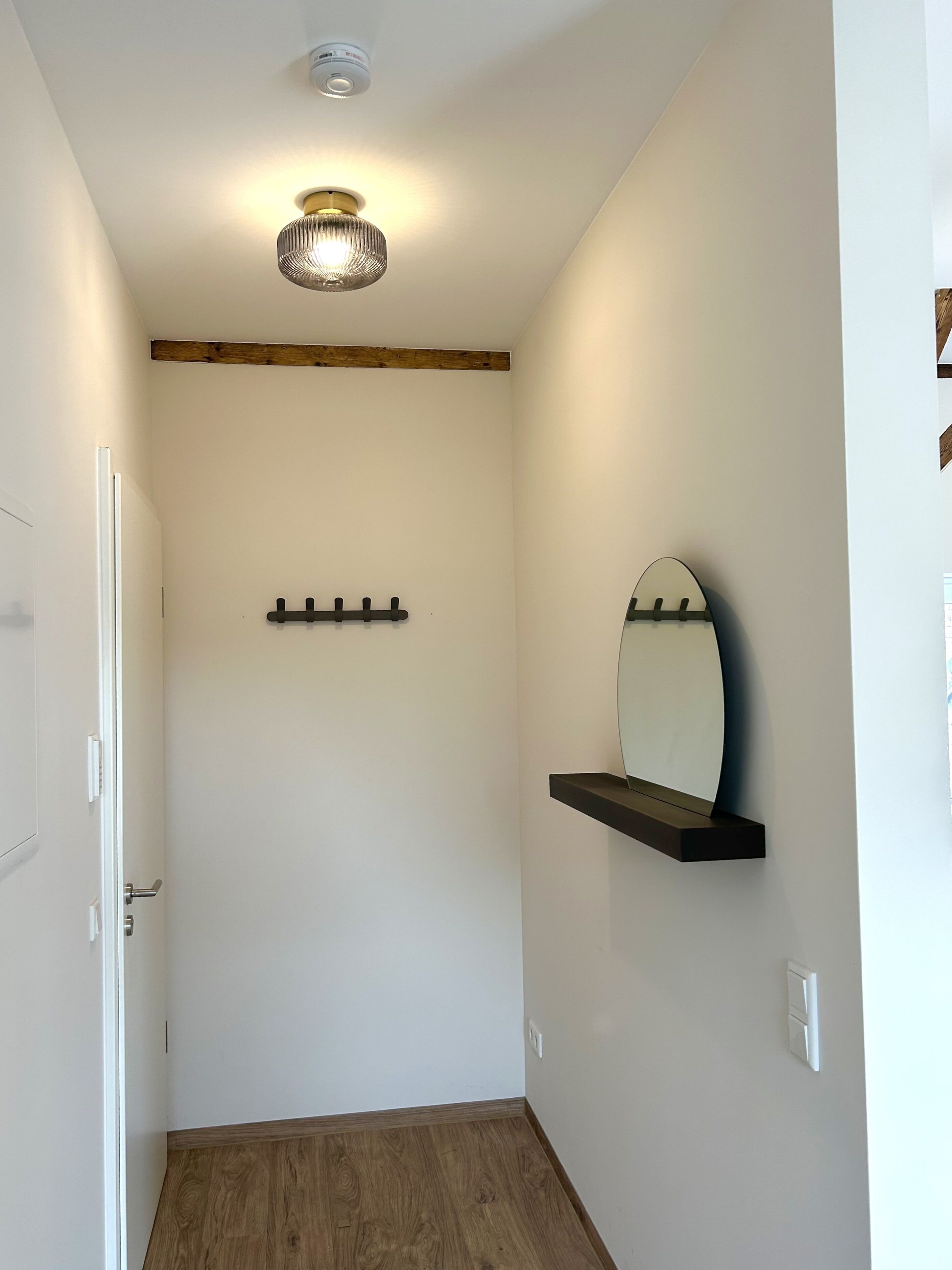 Passadís amb una porta de fusta, un llum al sostre, una estantanya amb mirall penjada a la paret i un sòl de fusta.