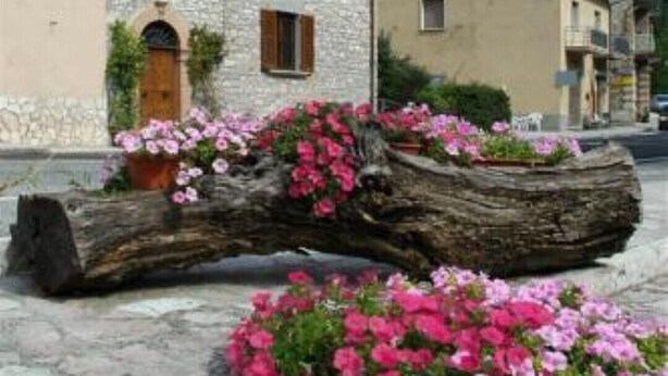 A stone building with a clock tower, surrounded by flower beds and a log. - Residence the garden on the Nera river - apt2 (Borgo Cerreto)