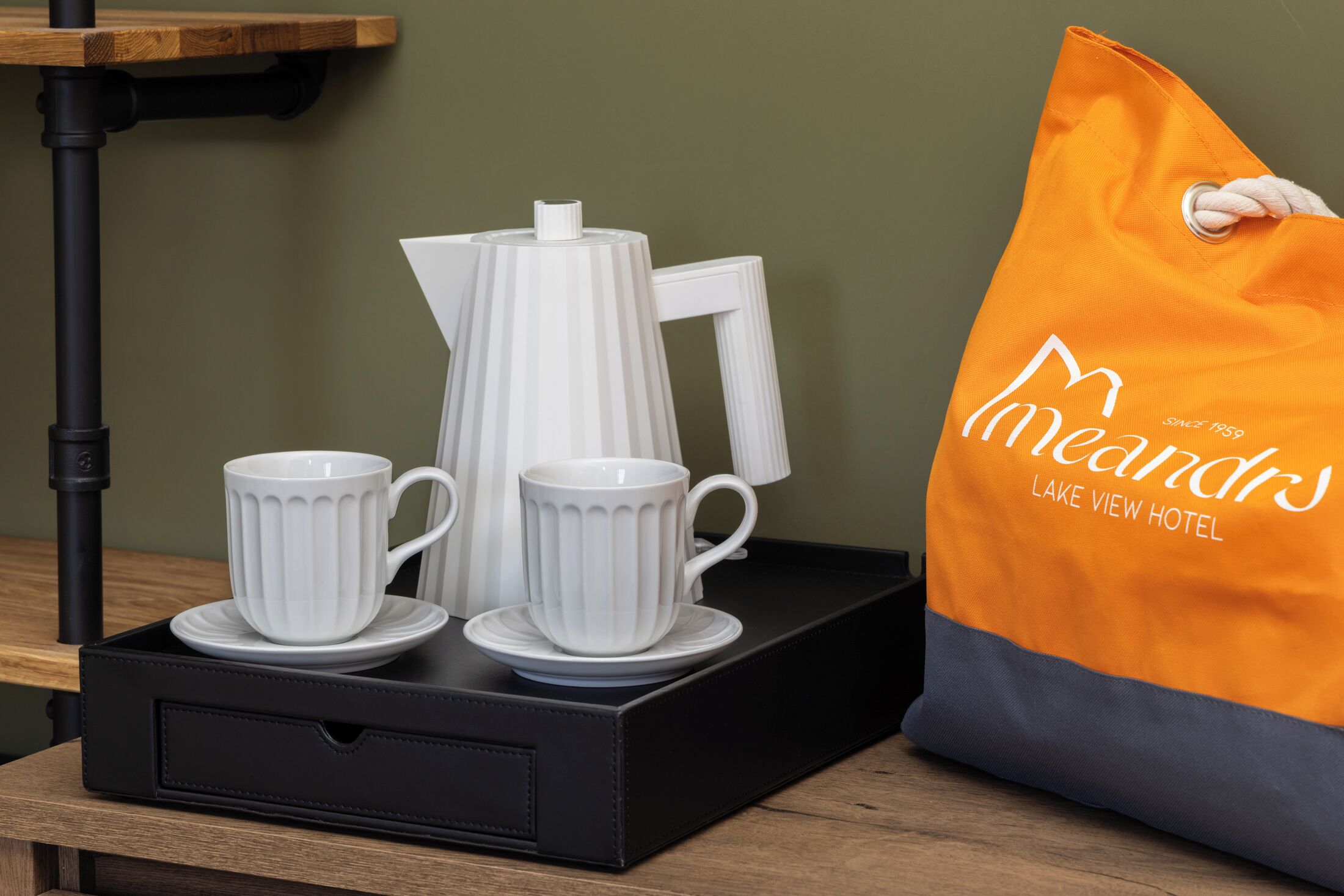 Una bandeja negra con dos tazas blancas y una tetera blanca, junto a un bolso naranja con texto.