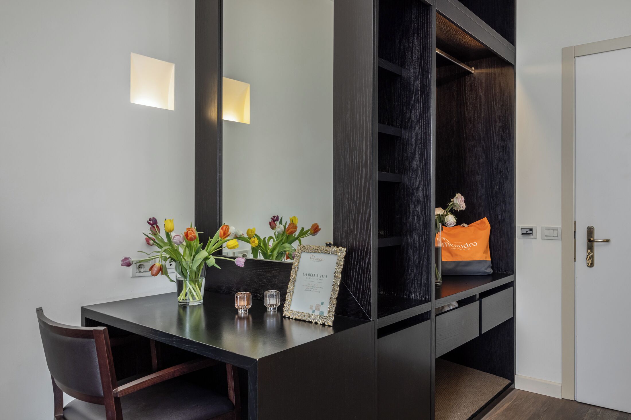 Una habitación moderna con un escritorio de madera oscura, un jarrón de tulipanes, un certificado enmarcado y un pequeño bolso naranja.