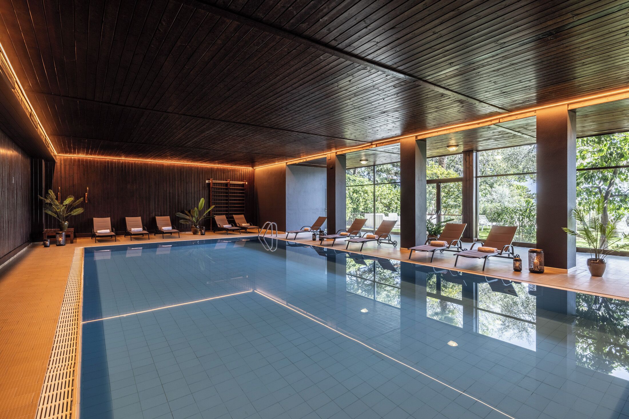 An indoor swimming pool with wooden ceiling and walls, sunlit windows, and lounge chairs along the poolside.