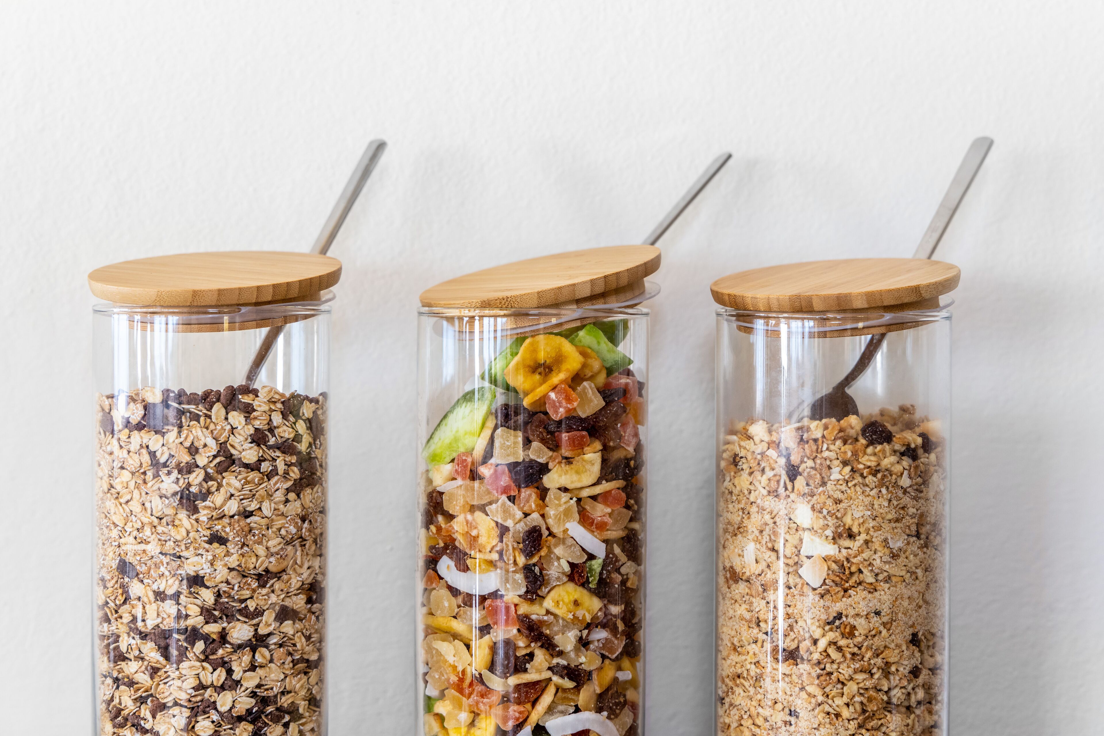 Three glass jars with wooden lids and metal straws, containing different types of granola.