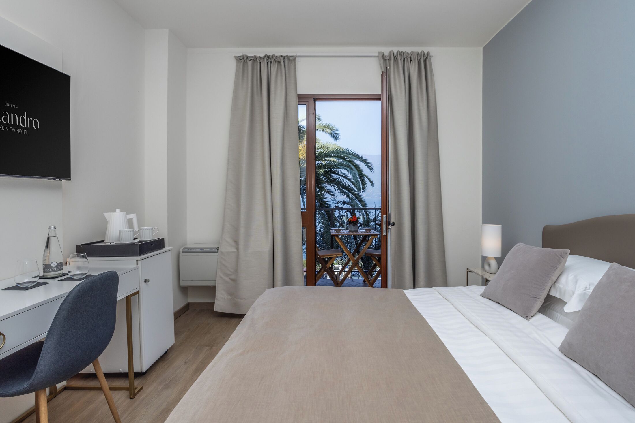 A hotel room with a bed, a desk, a chair, a TV, and a balcony with a view of palm trees.