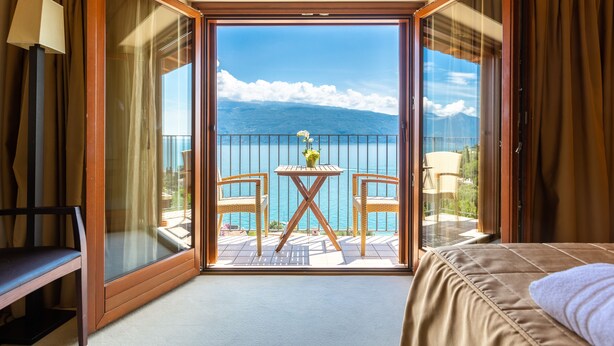 A hotel room with a balcony, a bed, a table with chairs, and a view of the sea and mountains. - Hotel Meandro - Lake View (Gargnano)