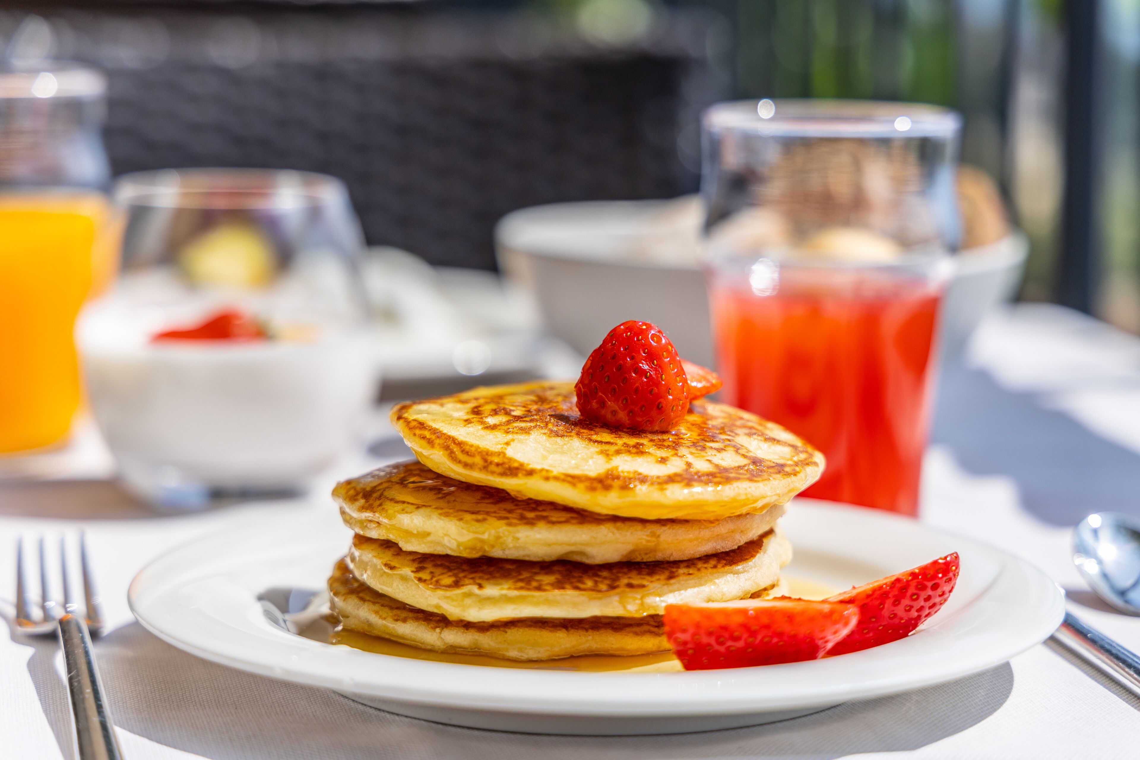 Una pila de tortitas en un plato con una fresa encima, acompañada de un vaso de jugo rojo y un vaso de jugo de naranja al fondo.