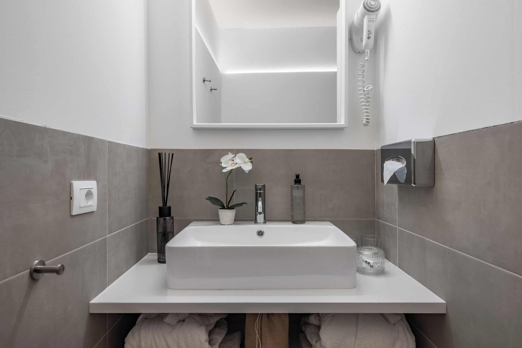 A modern bathroom with a white sink, a mirror, a hairdryer, a potted orchid, and a glass candle holder.