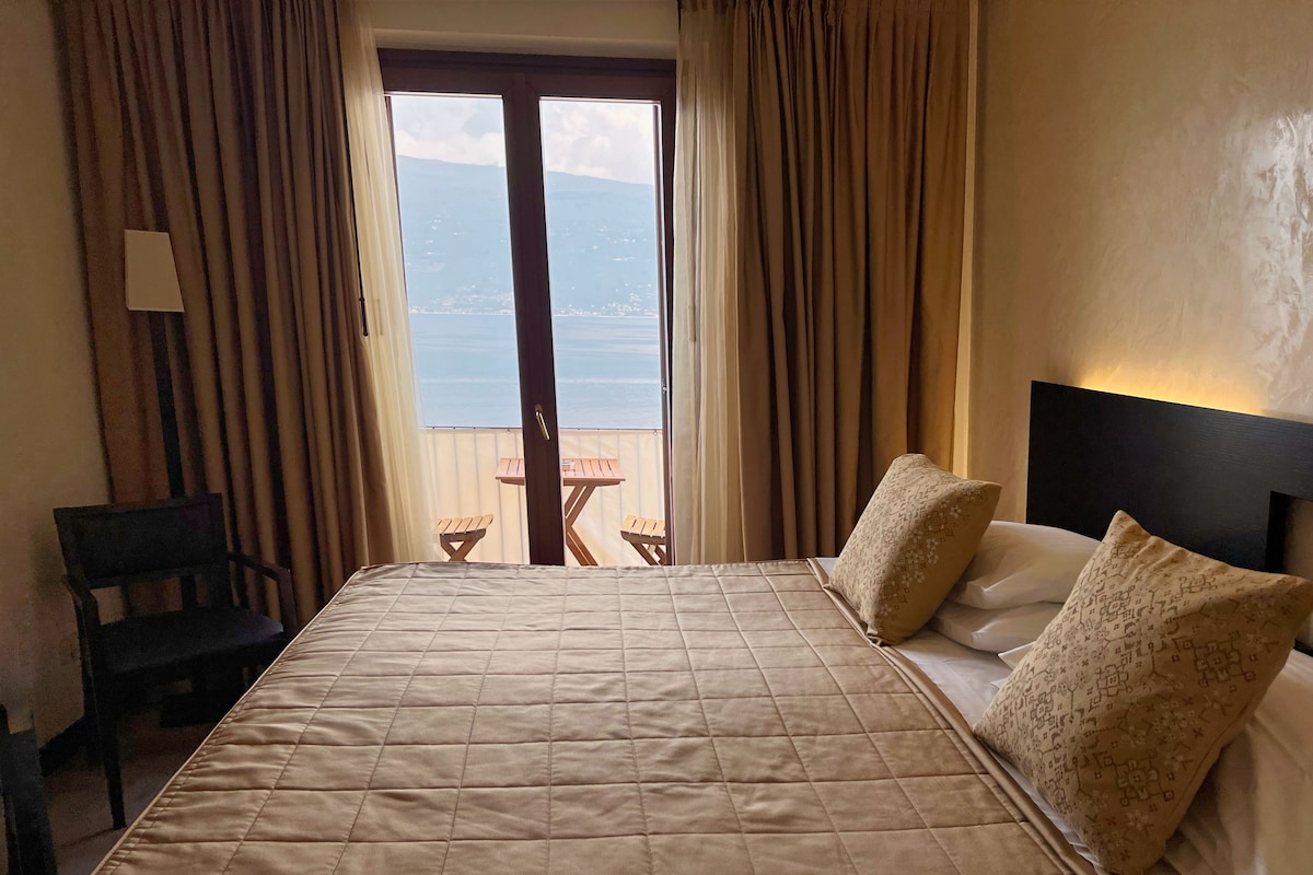 Habitación de hotel con una cama, dos almohadas, una silla y vista al mar a través de una ventana.