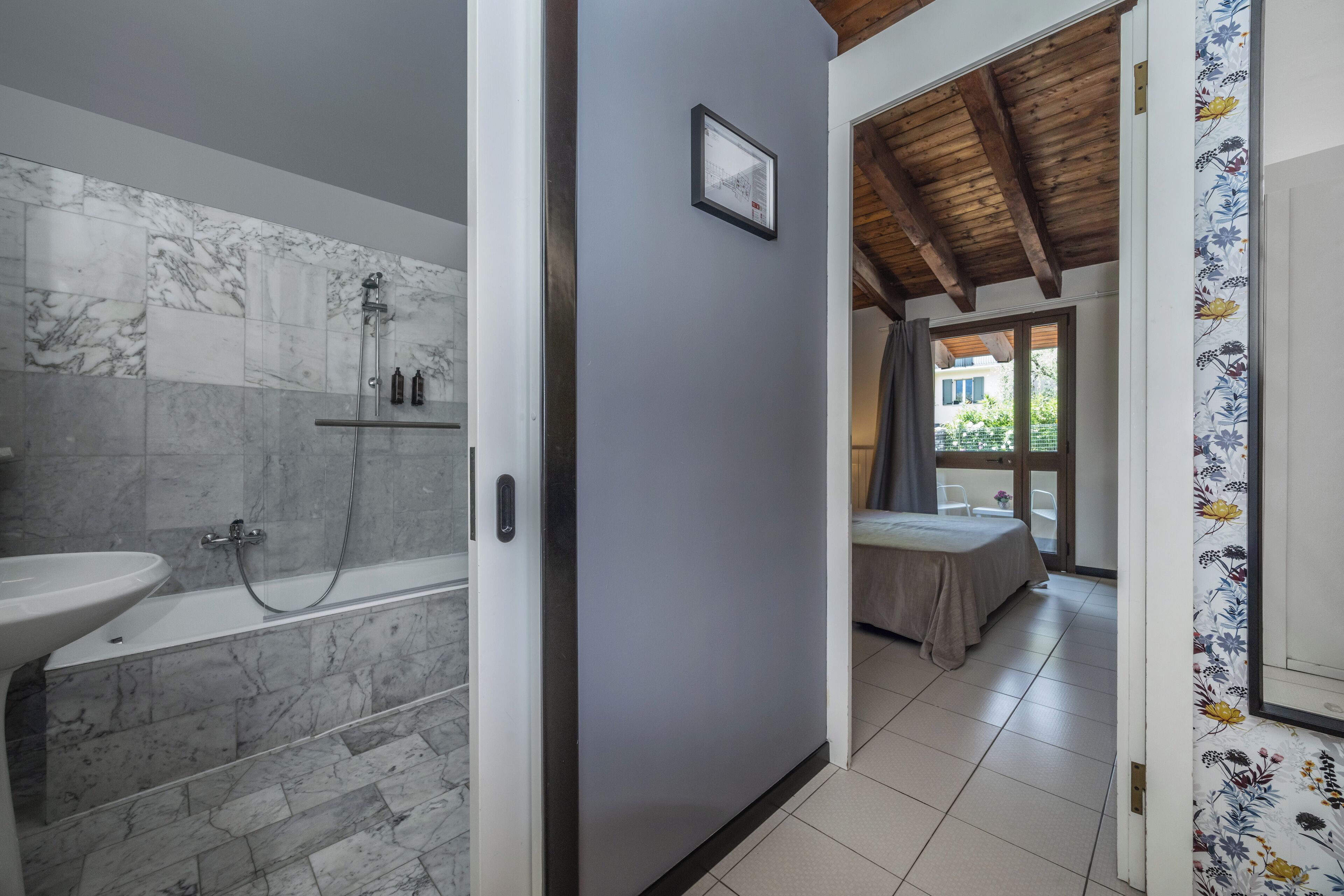 Un baño moderno con ducha de mármol, un lavabo y una bañera. Se ve una cama a través de una puerta abierta en la habitación contigua.