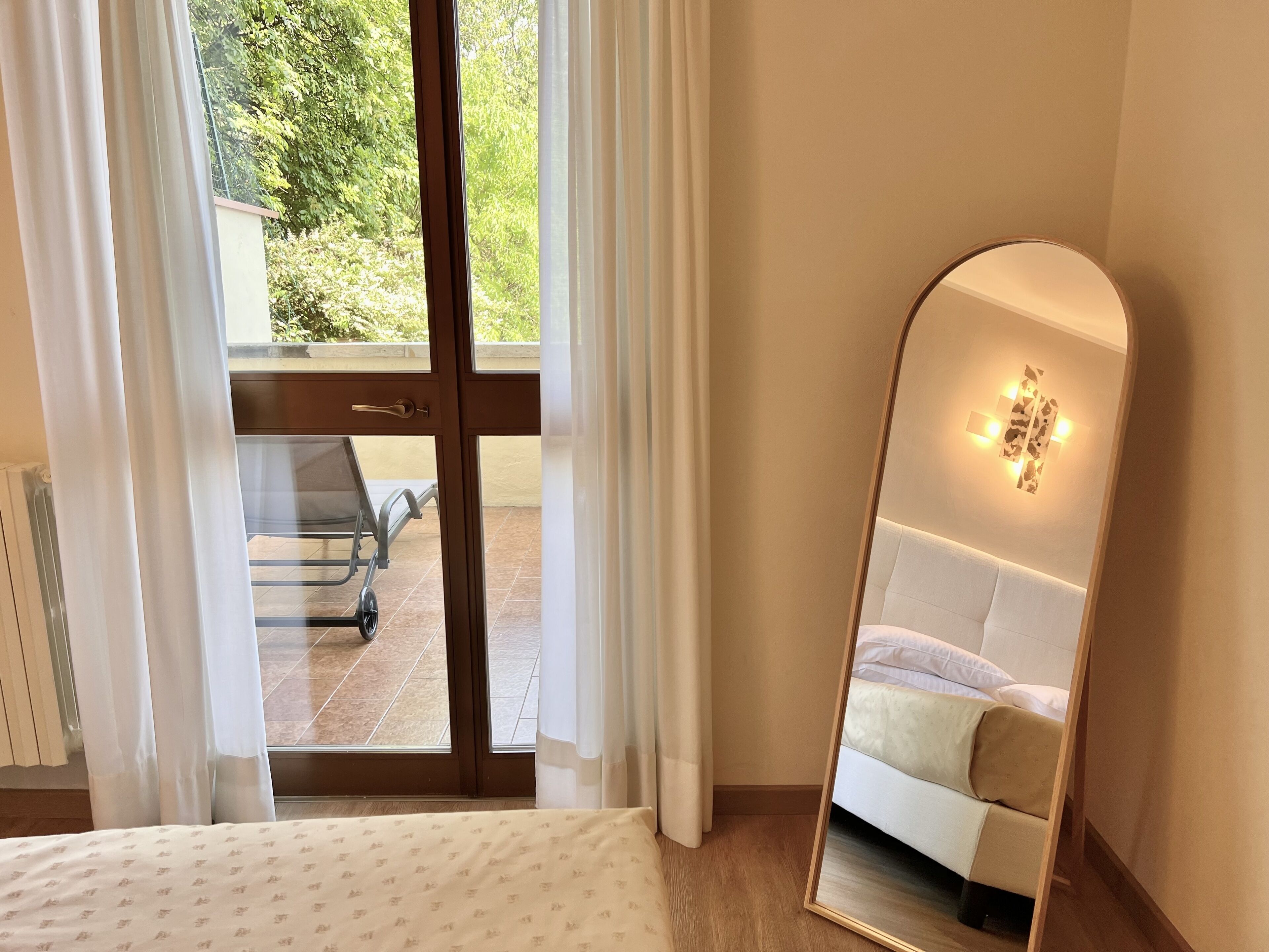 Ein Schlafzimmer mit Bett, einem großen Spiegel und einer Schiebeglastür, die zu einem Außenbereich mit Stuhl und Tisch führt.