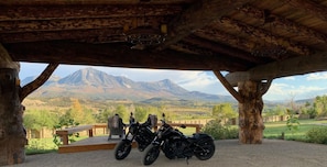 A covered outdoor seating area with two motorcycles parked under a wooden structure, overlooking a mountainous landscape.