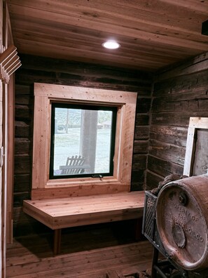 A cabin interior with a wooden bench under a window, a metal barrel, and a wooden ceiling. - Macy's Muse Log Cabin (Paonia)