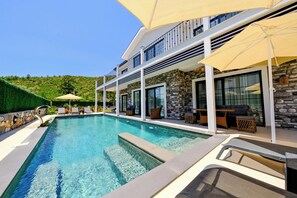 Una piscina con sillas de descanso y sombrillas, un muro de piedra y un edificio moderno con balcones.