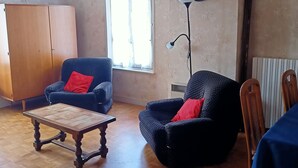 Ein Wohnzimmer mit Holzboden, zwei Sesseln, einem kleinen Tisch, einem Deckenventilator, einem Fenster mit Vorhängen und einer Heizung.