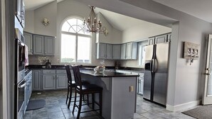 A spacious kitchen with grey cabinets, a large island with bar stools, a stainless steel refrigerator, and a chandelier.