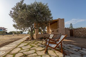 A stone patio with two wooden lounge chairs, a stone wall, and a tree. - Beachfronthouse in Aegina (Aegina)
