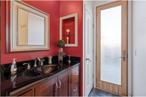 A bathroom with a red wall, a large mirror, a sink with a black countertop, and a wooden door with frosted glass. - Restful Retreat in CaveCreek, N Scottsdale (Cave Creek)