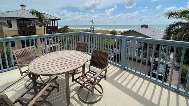 A balcony with a wooden table and chairs, overlooking a beach with a clear sky. - Florida Beachfront Bliss just steps from the Sand (St. Pete Beach)