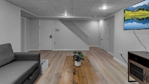 A modern living room with a grey sofa, wooden flooring, a staircase, and a TV mounted on the wall. - Modern Trendy Downtown (Buffalo)