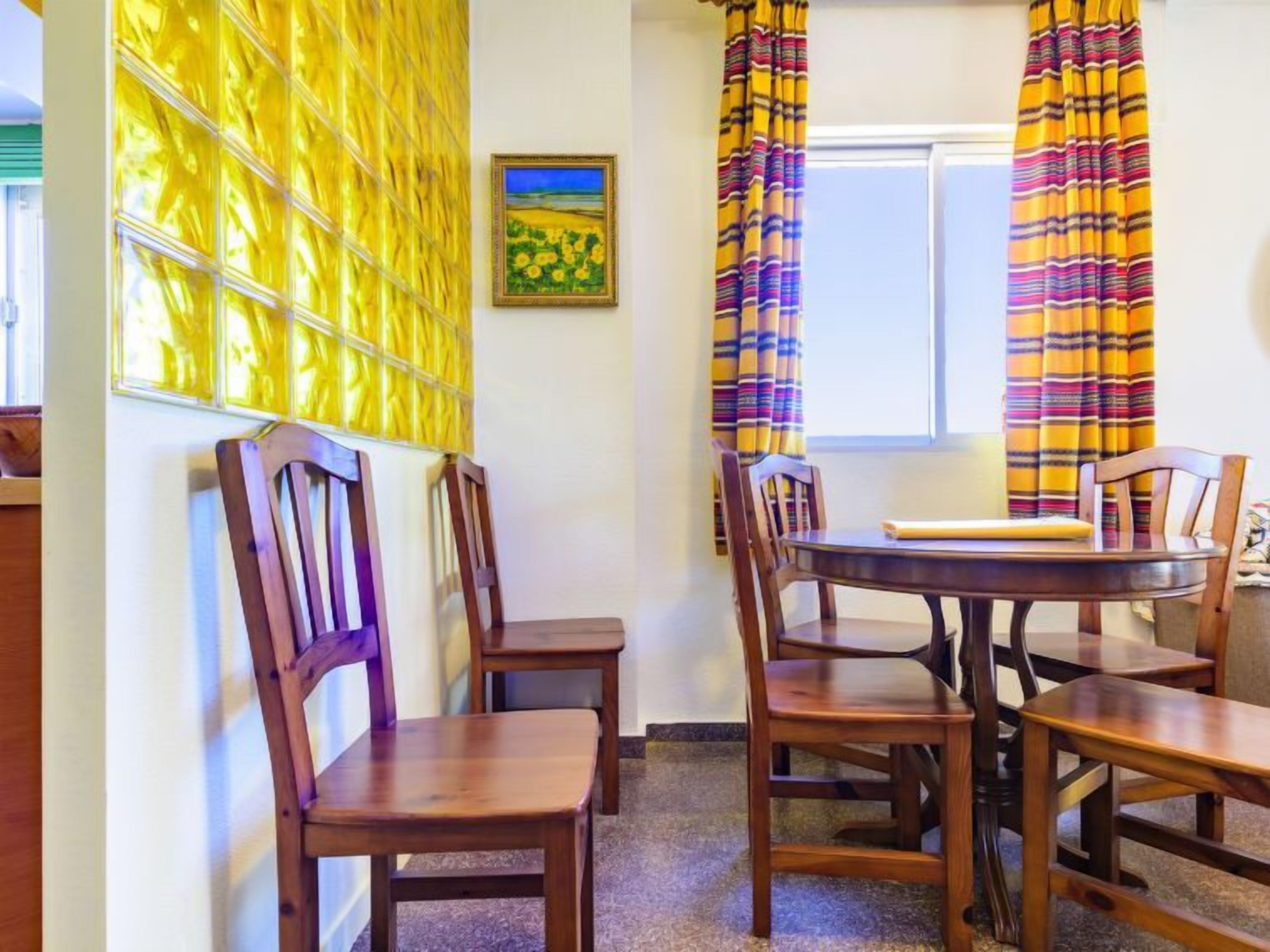Un comedor con sillas de madera y una mesa, cortinas amarillas y un cuadro en la pared.