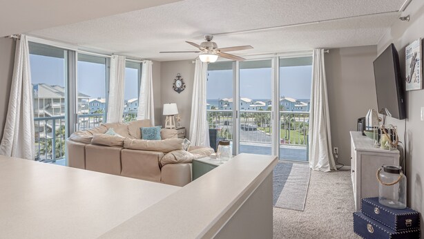 A spacious living area with a beige sofa, a ceiling fan, and large windows offering a view of the sea and other buildings. - Gulf & Bay Views: Sleeps 6 in 3BR, 2BA Condo-TT3F (Pensacola Beach)