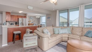 A modern living room with a beige sectional sofa, a wooden kitchen with a bar area, and large windows offering a view of the neighborhood. - Gulf & Bay Views: Sleeps 6 in 3BR, 2BA Condo-TT3F (Pensacola Beach)