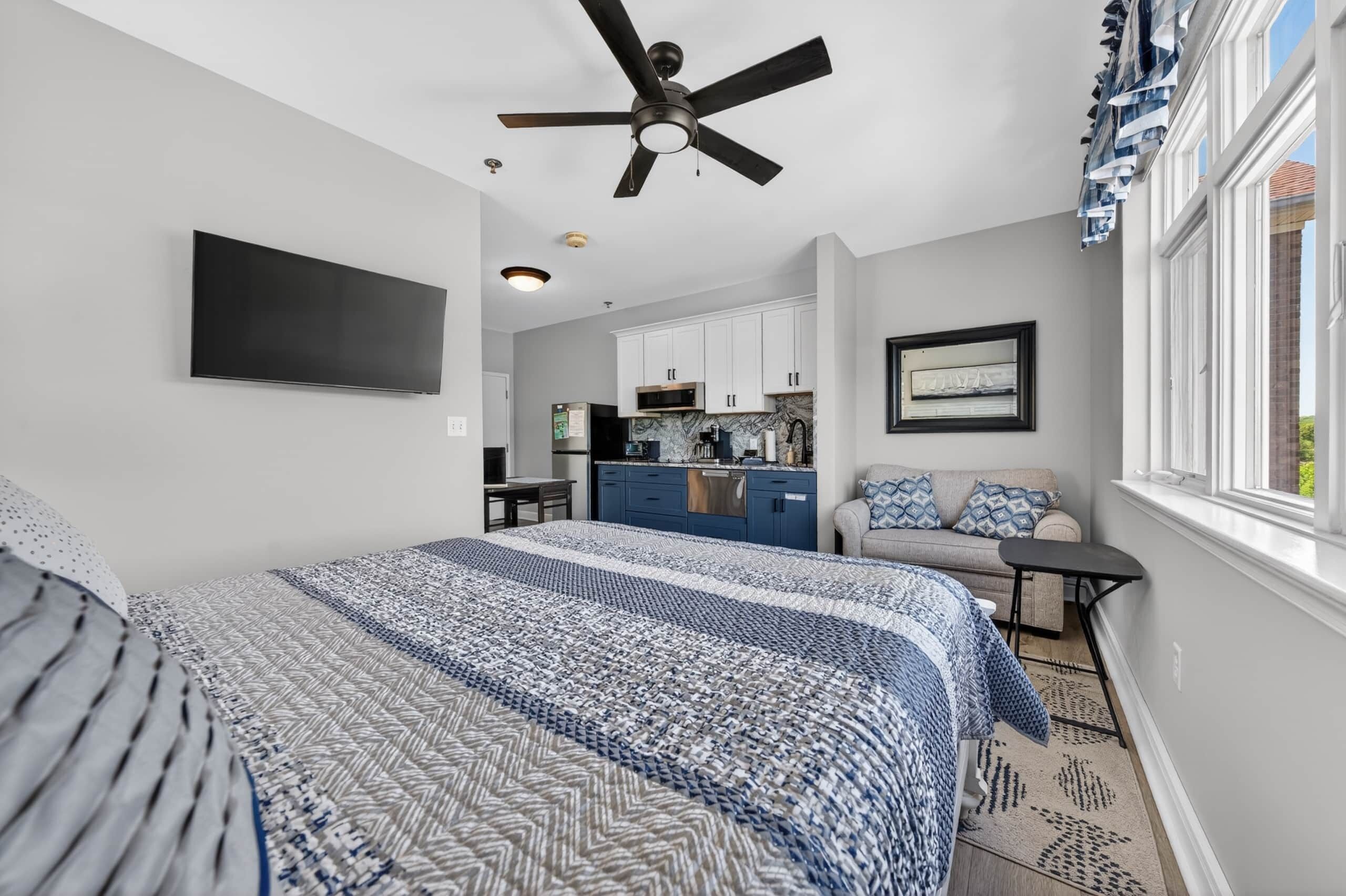 A bedroom with a bed, a ceiling fan, a TV, a kitchenette, and a window.