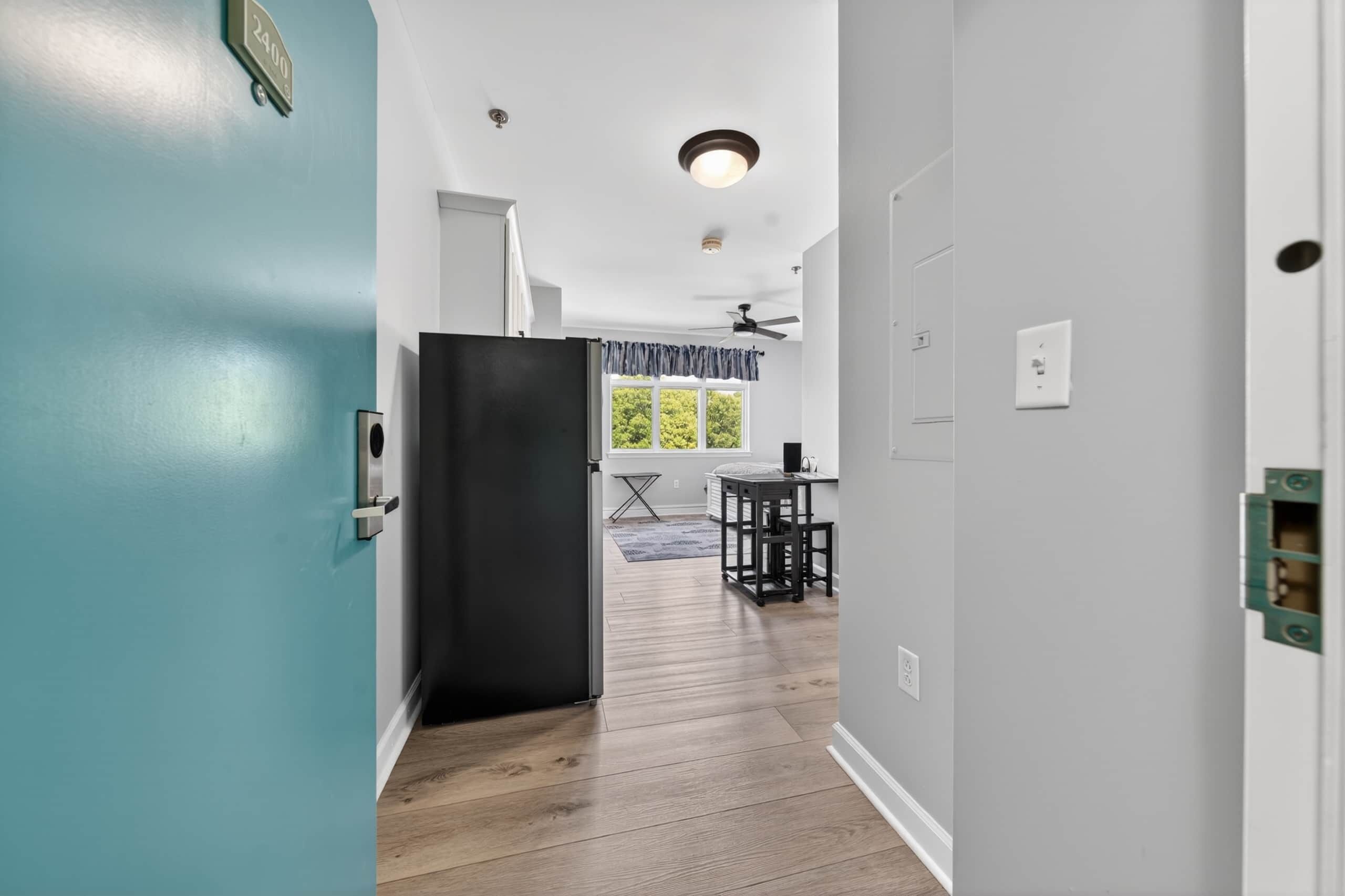 Un couloir avec une porte de couleur turquoise, un réfrigérateur noir, un coin repas avec une table et des chaises, et un ventilateur de plafond.