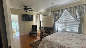 A bedroom with a bed, a desk, a chair, a ceiling fan, and a TV mounted on the wall.