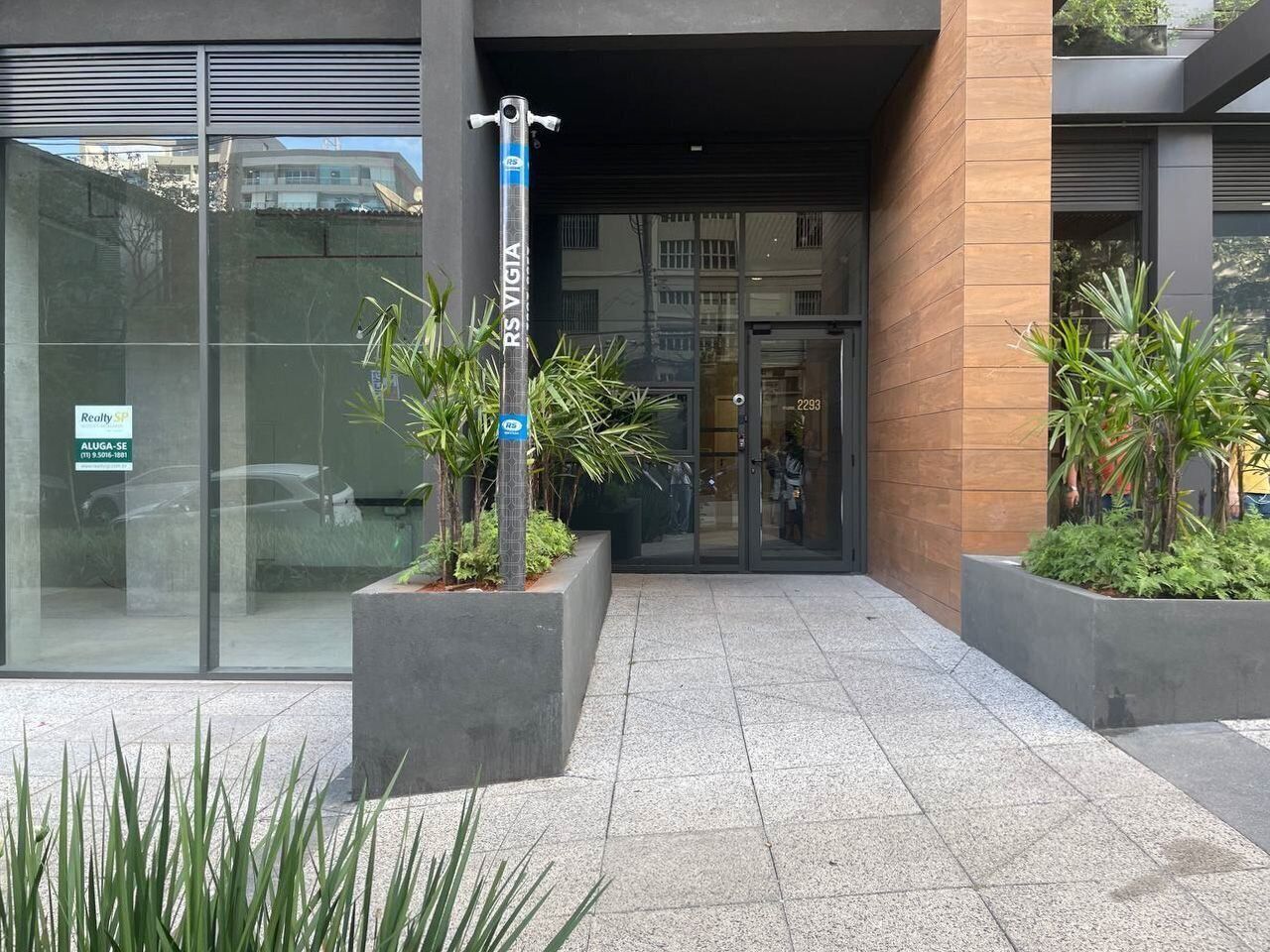 A modern building entrance with a glass facade, a metal pole with signage, and potted plants.
