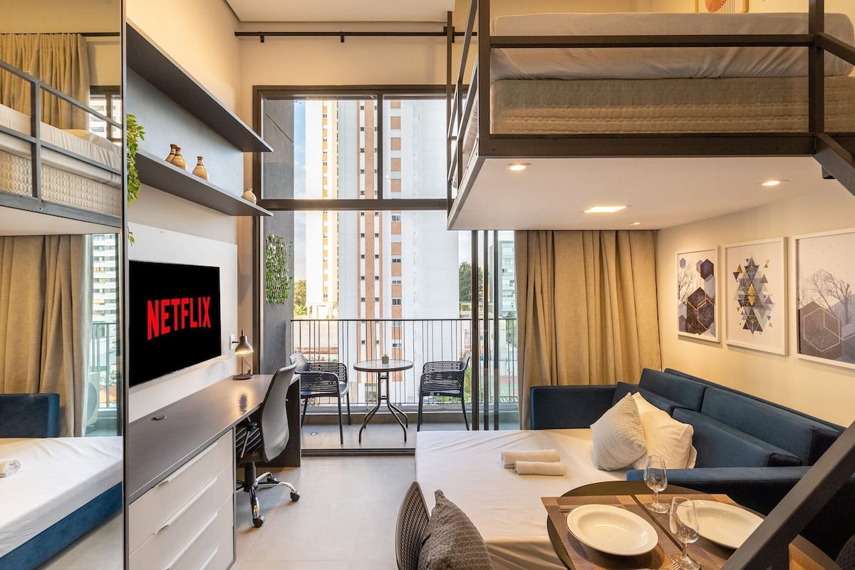 Quarto de hotel moderno com varanda, cama, sofá, escrivaninha e televisão exibindo o logotipo da Netflix.