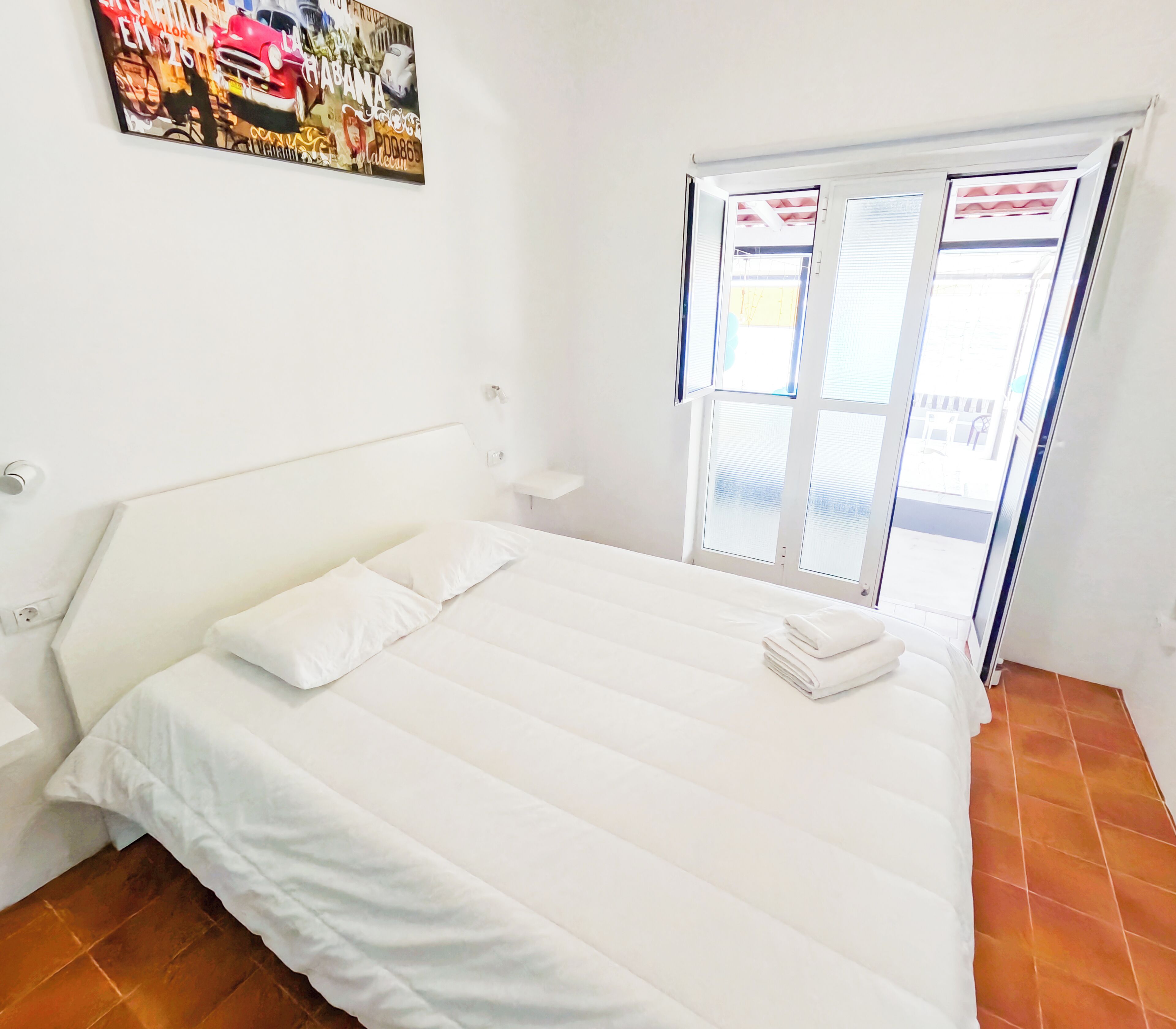 Uma cama grande com roupa de cama branca, uma mesinha de cabeceira e uma janela com persianas.