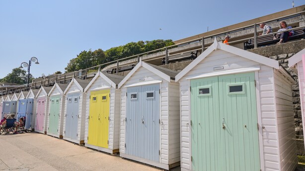 Eine Reihe bunter Strandhäuser mit weißen Fassaden und pastellfarbenen Türen, angeordnet entlang einer Strandpromenade.