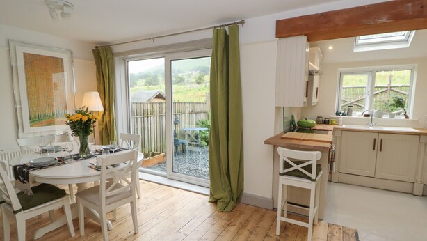 A dining area with a table and chairs, a kitchen with cabinets and a sink, and a sliding glass door leading to a garden. - Beech Croft (Keswick)