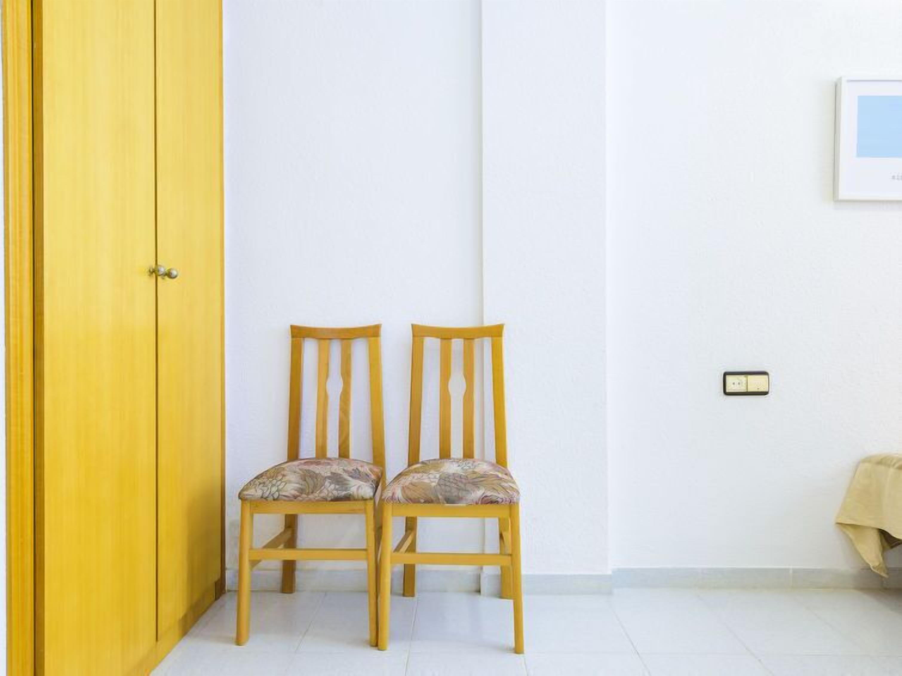 Una habitación con una puerta amarilla, dos sillas de madera con cojines florales, una pared blanca y un pequeño interruptor de luz.
