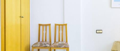 Una habitación con una puerta amarilla, dos sillas de madera con cojines florales, una pared blanca y un pequeño interruptor de luz.