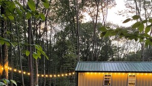 Una cabaña rústica con techo de metal, rodeada por un sendero de grava y escalones de piedra, ubicada en un área boscosa con guirnaldas de luces.