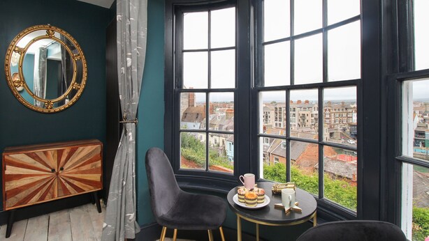 Une pièce dotée d’une grande baie vitrée, d’un miroir rond, d’une petite table avec des pâtisseries et une tasse, et d’un fauteuil de couleur sombre.