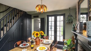Un coin repas avec une table en bois dressée pour quatre personnes, entourée de chaises en cuivre. Un vase rempli de tournesols est posé sur la table, et une bouteille de vin est visible.