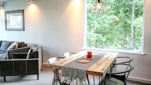 Un espace repas moderne avec une table en bois, des chaises en métal, un chemin de table à motifs et une grande fenêtre donnant sur la forêt.