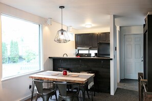 Enjoy meals in this cozy kitchen with a dining table for two, a modern range hood, and a large window offering a view of the outdoors.
