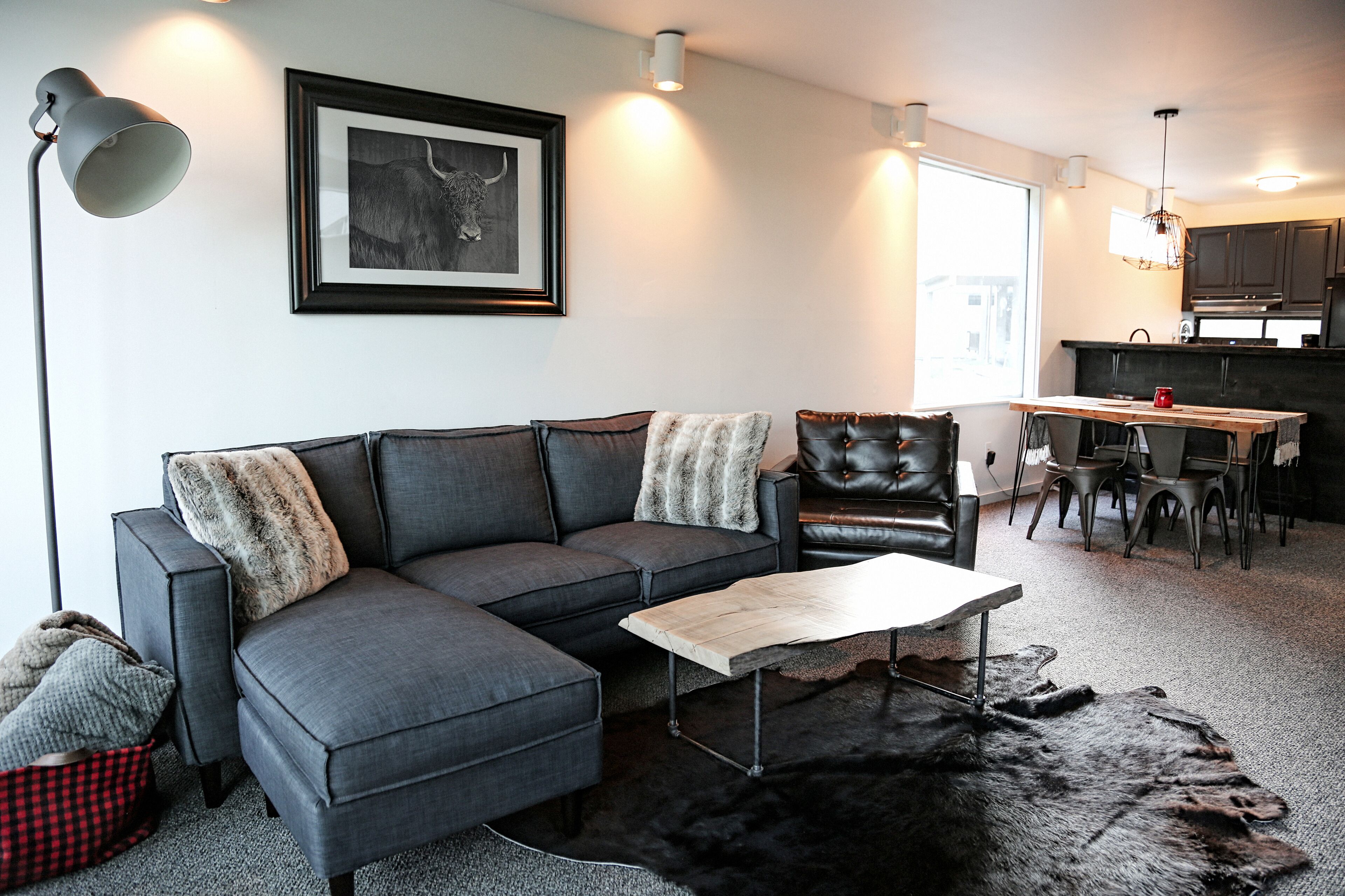 Un salon moderne avec un canapé d’angle gris, une table basse en bois et un fauteuil en cuir noir. Un tableau encadré orne le mur, on aperçoit un coin repas avec une tasse rouge sur la table, et une cuisine en arrière-plan.